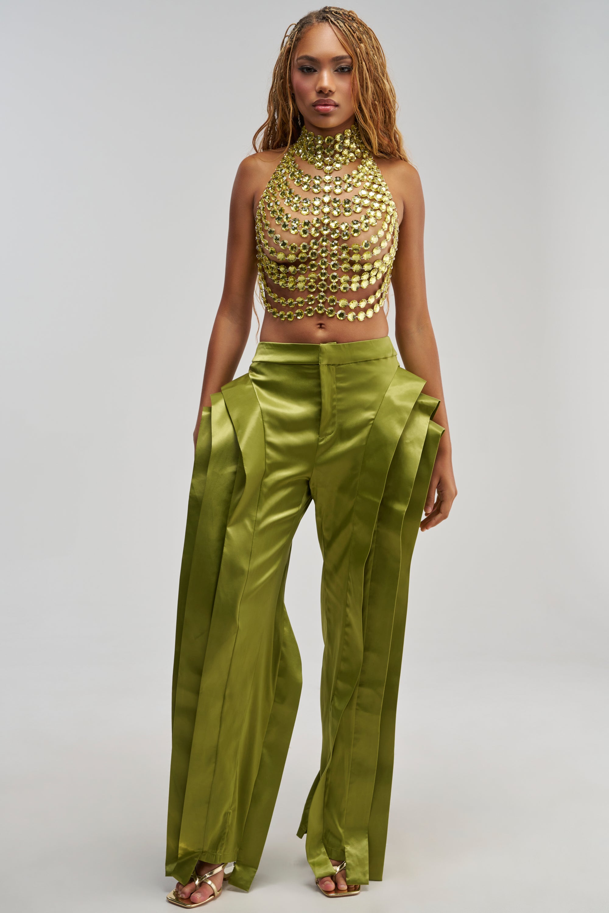 A woman stands against a plain light-grey background wearing the INFINITY TROUSER IN OLIVE—shiny satin trousers with a high-rise fit and layered panels—paired with a gold metallic halter top.