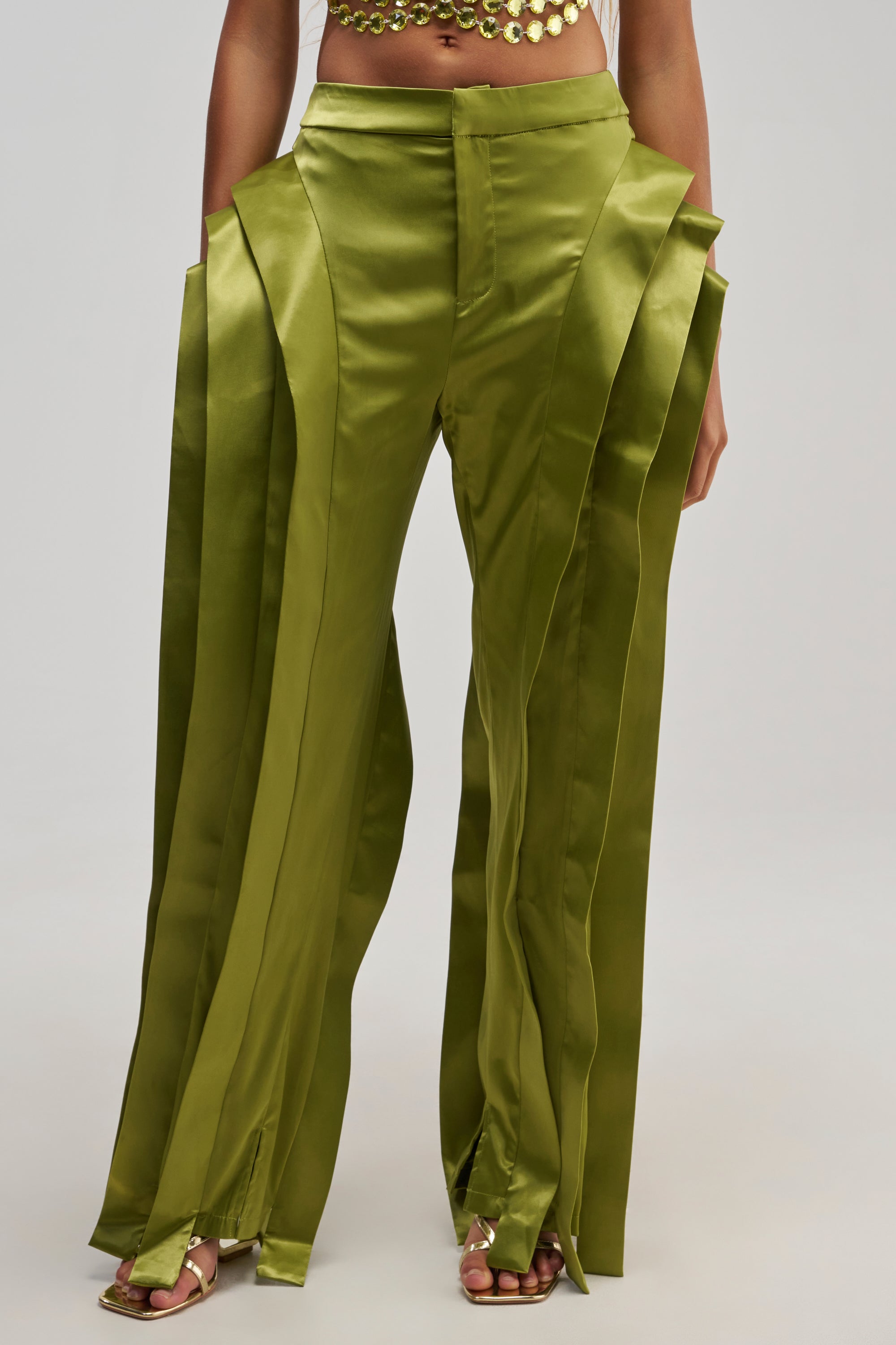 A woman is seen wearing the INFINITY TROUSER IN OLIVE—shiny green pleated pants featuring a high-rise fit and distinctive layered panels—with metallic sandals. The straight-leg silhouette of these satin trousers lends an air of sophistication. The photo is cropped at her midriff.