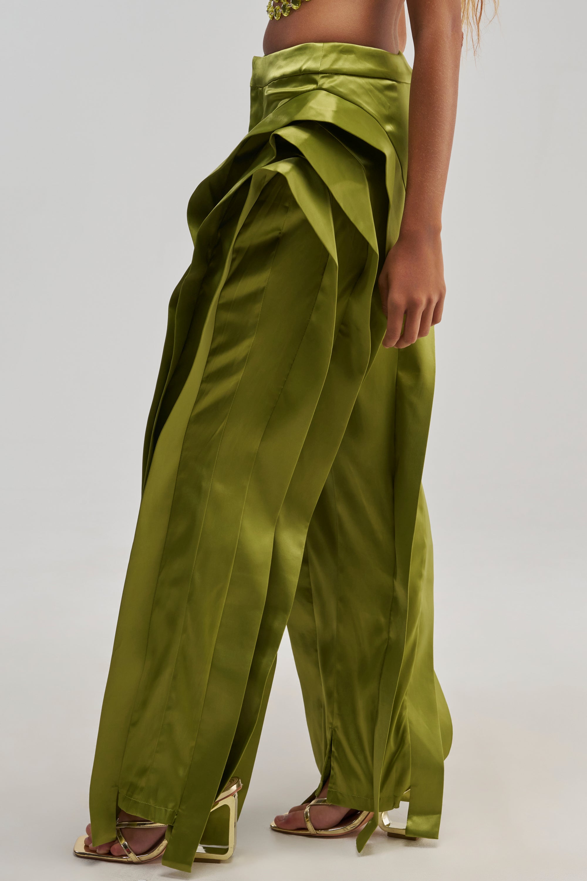 A person is wearing the INFINITY TROUSER IN OLIVE—shiny, satin wide-leg pants featuring layered, ribbon-like fabric strips cascading down the sides. The outfit is styled with open-toe heels, and only the lower body is shown against a light background.