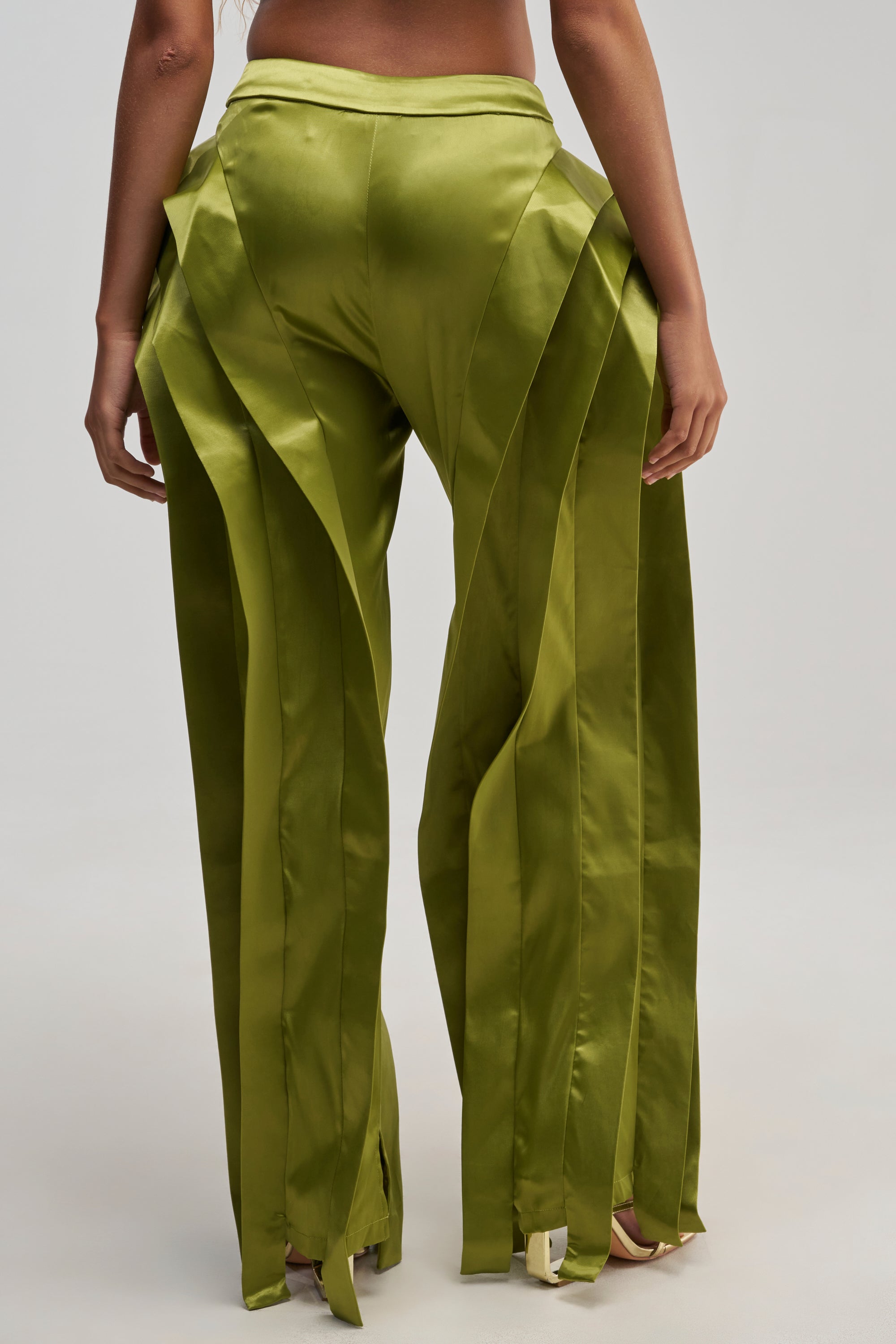 A person stands facing away, wearing the INFINITY TROUSER IN OLIVE—shiny, olive satin pants featuring layered, sculptural panels and a glossy texture that enhances their straight-leg silhouette.