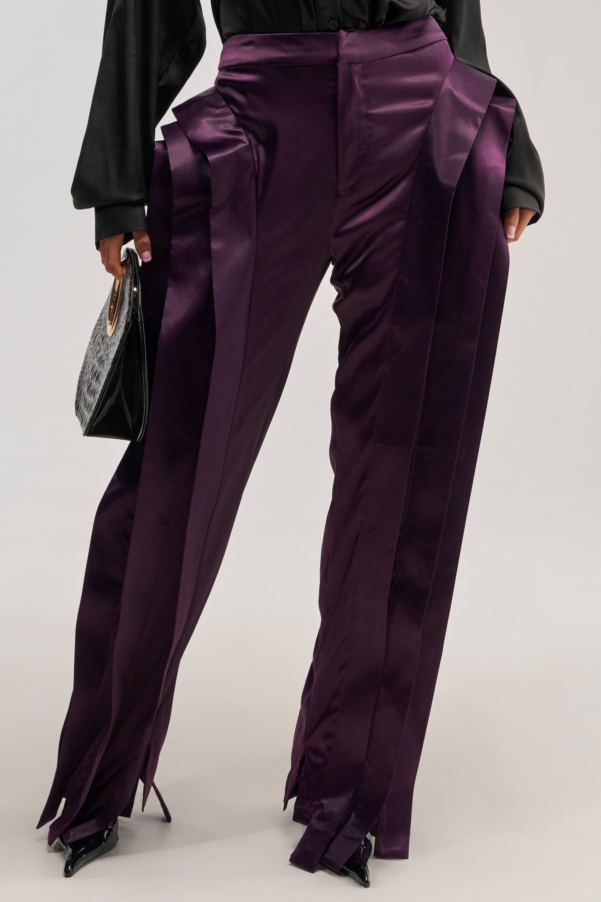 A person wears the INFINITY TROUSER in shiny purple satin featuring a high-rise fit with wide, pleated panels, paired with black pointed heels and a textured black handbag. The look is completed with a loose-fitting black blouse that complements the avant-garde silhouette, set against a plain light-colored background.
