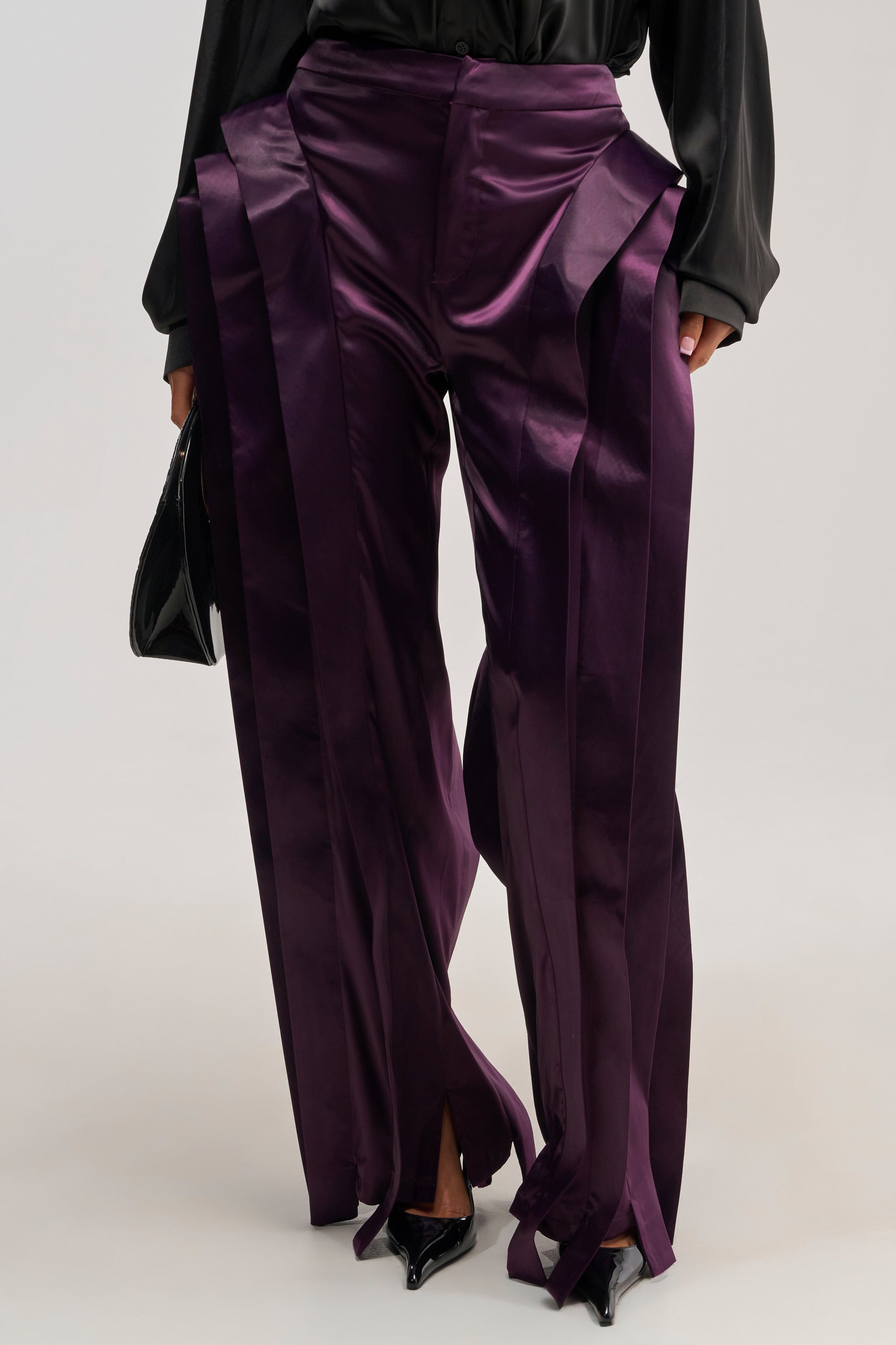 A person wearing the INFINITY TROUSER IN PLUM—shiny, deep purple, wide-leg pants with vertical pleats and a high-rise fit—paired with a black blouse, black pointed heels, and holding a black bag. The photo is cropped from the shoulders down.
