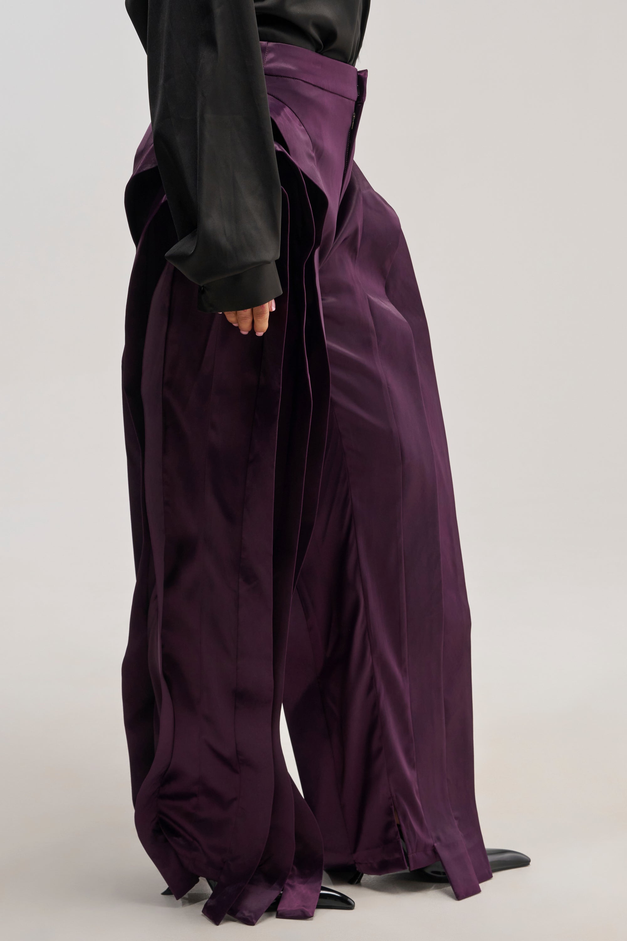 A person wearing the INFINITY TROUSER—featuring a wide-leg, high-rise fit in deep purple satin with layered panels—paired with a black long-sleeve top, standing against a plain background and showcasing an avant-garde silhouette.