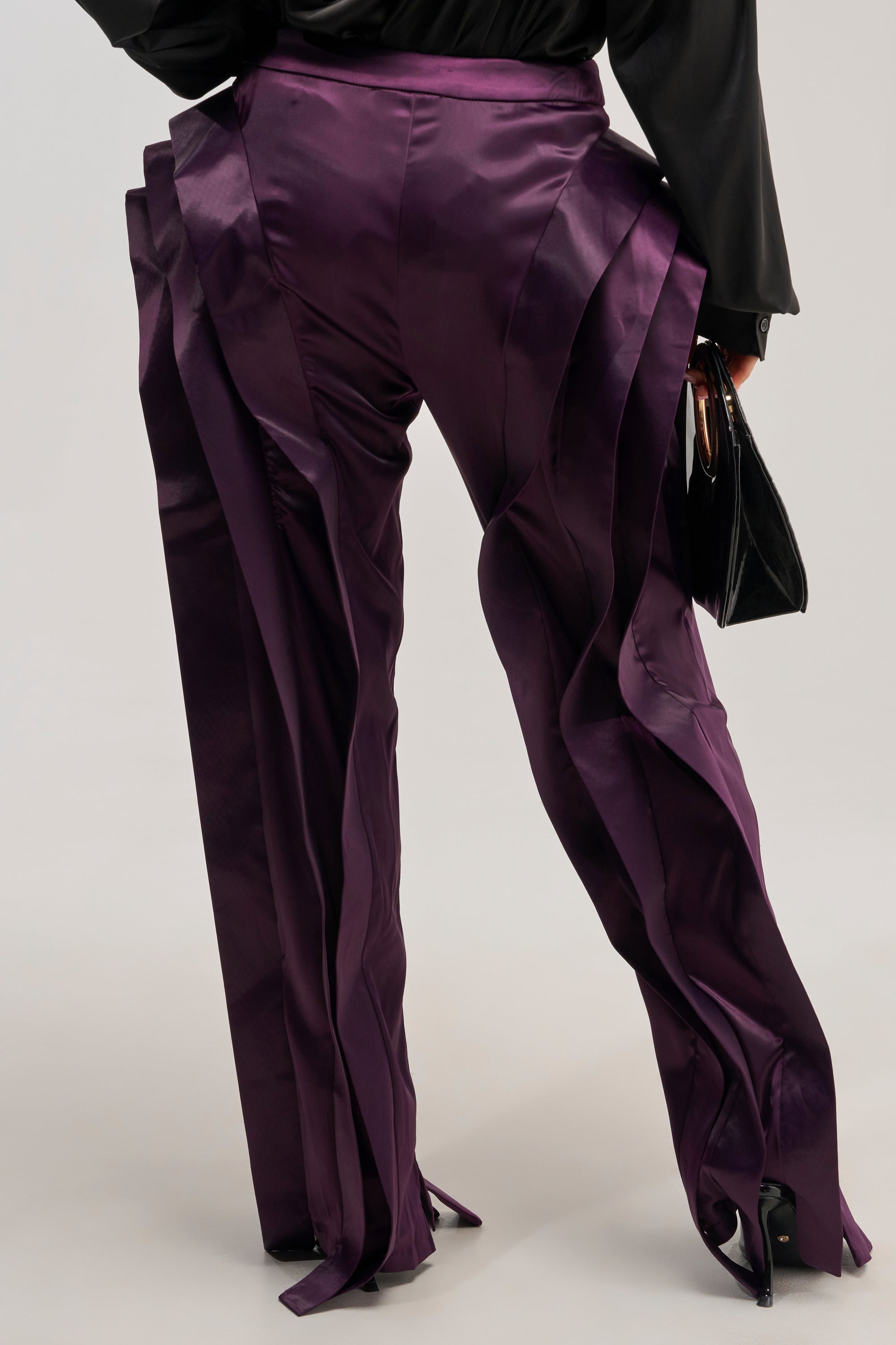 Wearing the INFINITY TROUSER in shiny purple satin, featuring dramatic layered ruffle detailing along the legs for an avant-garde silhouette. The trousers are styled with a black top, black shoes, and a small black handbag. The background is plain and light-colored.