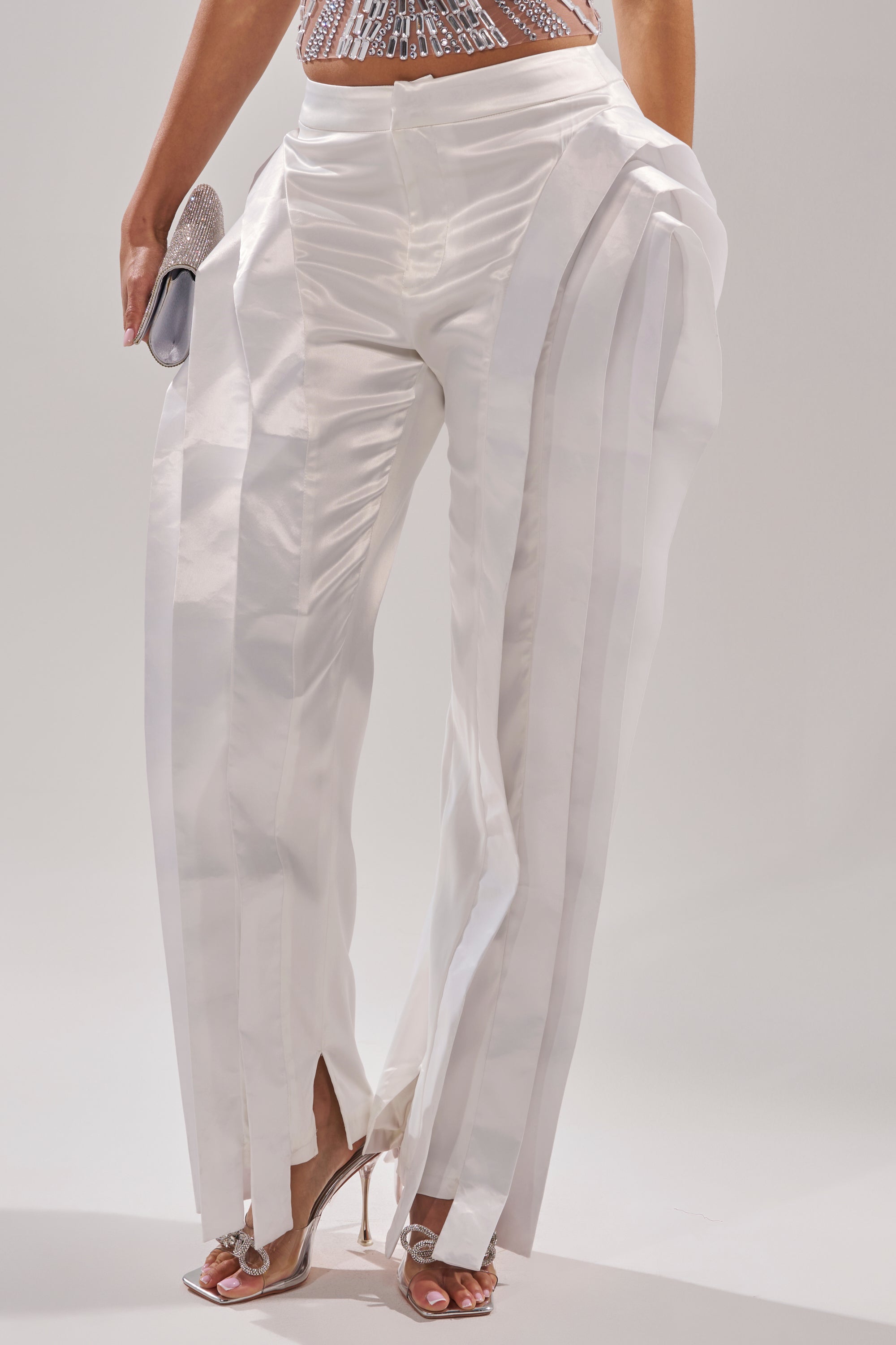 A person wearing the INFINITY TROUSER—shiny white high-rise straight-leg pants with vertical pleats and front ankle slits—paired with silver heels and a matching clutch. Only their lower half is shown.