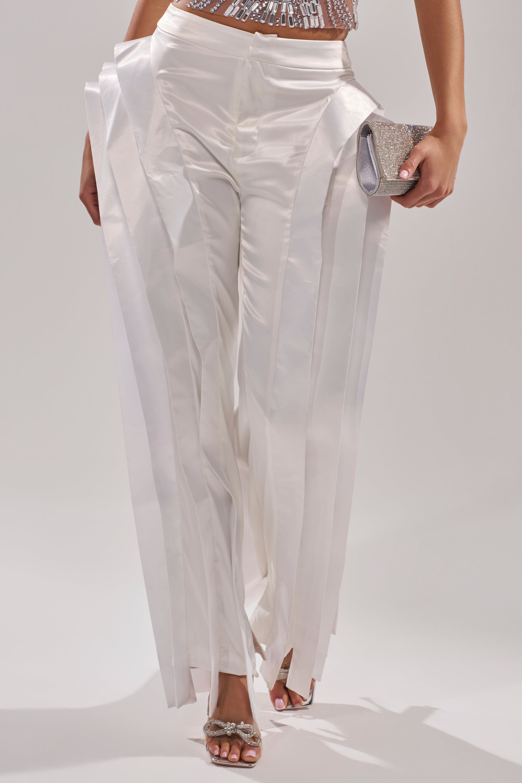 A person wears the INFINITY TROUSER in shiny white satin with vertical pleats, paired with a sequined top, silver heeled sandals, and a glittery silver clutch against a plain, light-colored background.