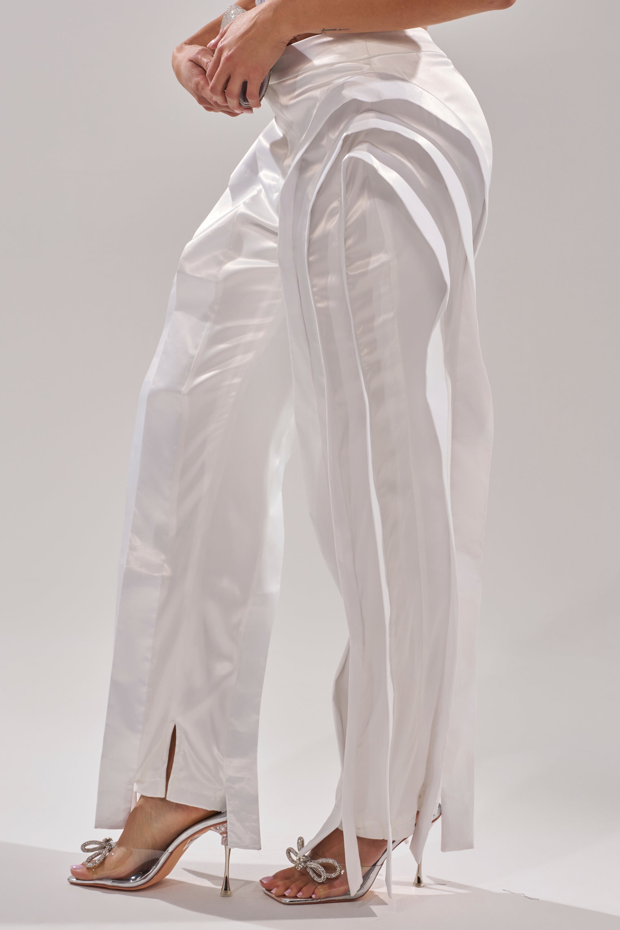 Here is a rewritten version of your sentence using the provided product data: The INFINITY TROUSER IN WHITE features a high-rise fit and wide-leg silhouette with dramatic draped panel details, styled for sophisticated evening wear with silver stiletto heels adorned with crystal bow embellishments, worn by a woman against a light grey background.