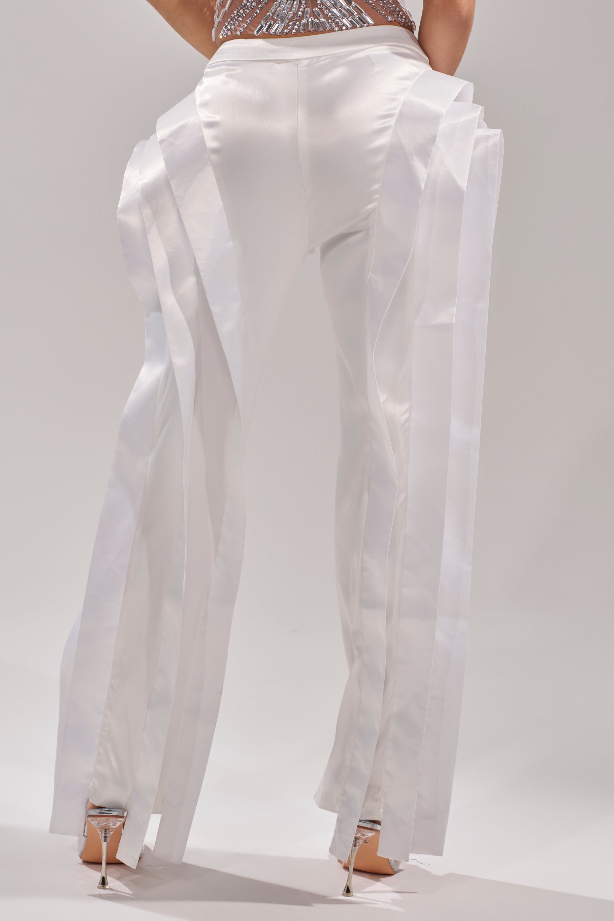A person in high heels and the INFINITY TROUSER—a high-rise, satin pant with shiny white panels—seen from behind against a plain, light background.