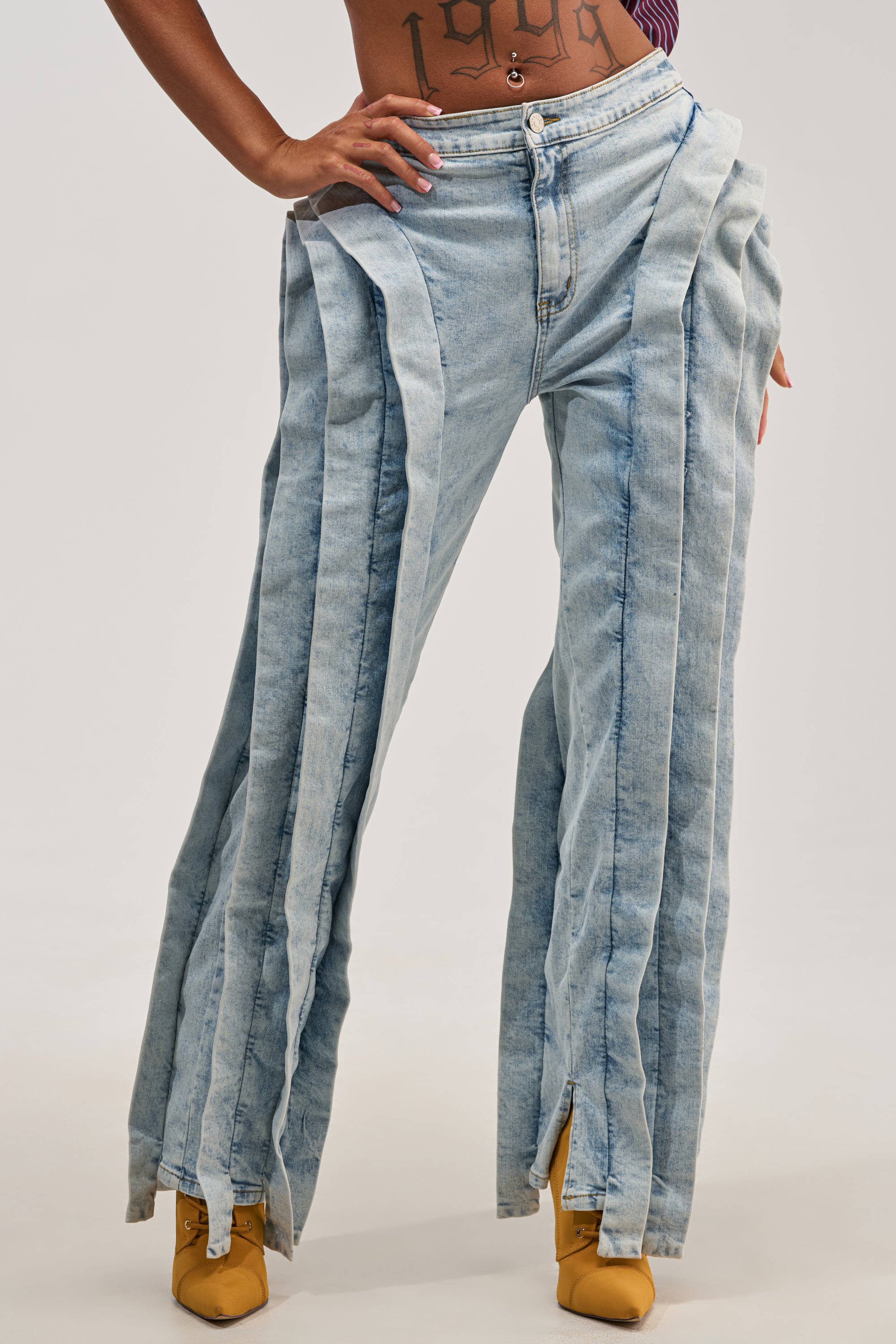 Wearing the INFINITY DENIM—light wash, high-waisted jeans featuring wide, pleated panels running down each leg—paired with yellow pointed-toe shoes, this look offers a fresh twist on avant-garde style. A tattooed midriff peeks out, lending an extra dose of bold streetwear attitude.