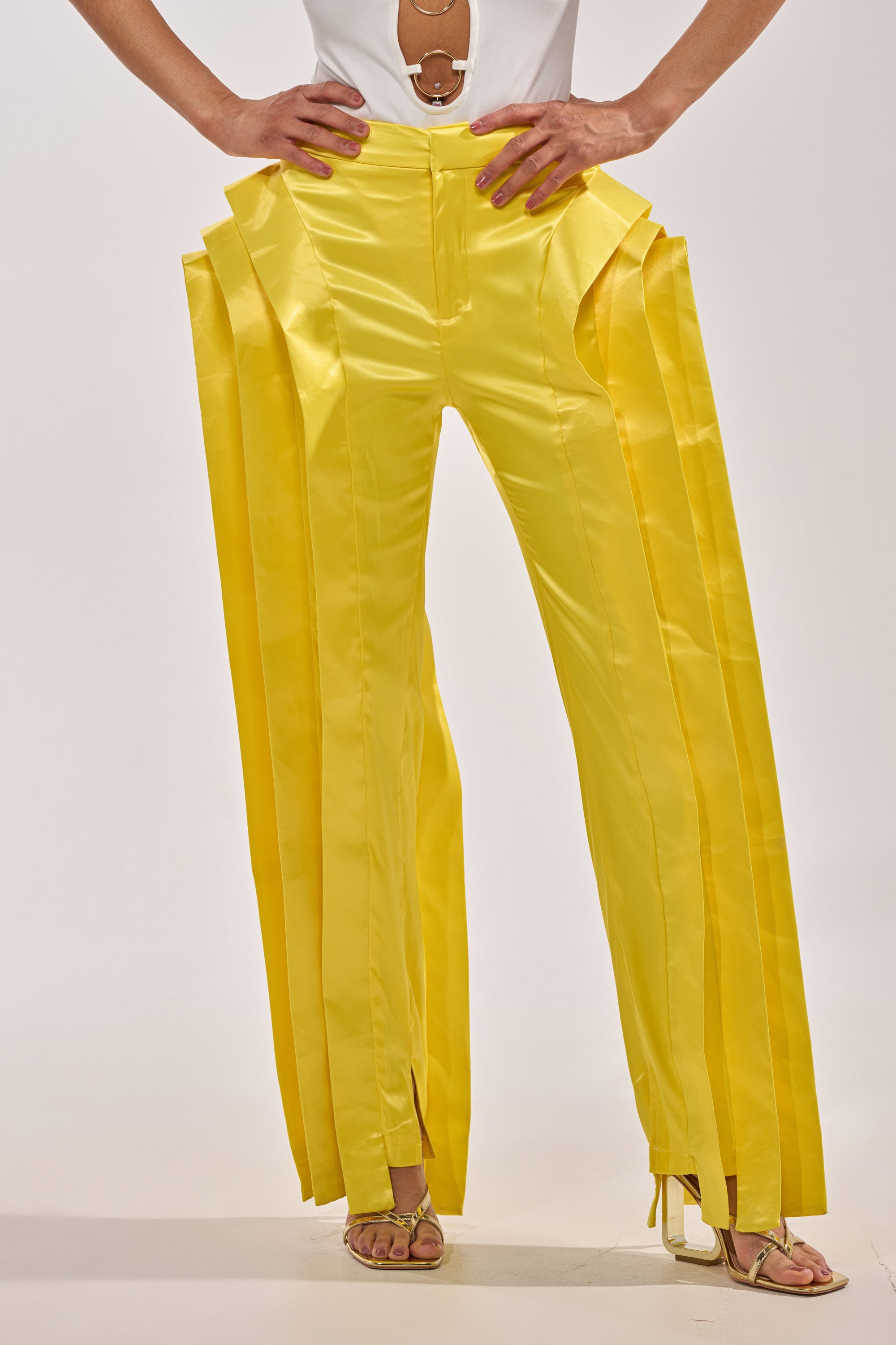 Someone models the YELLOW INFINITY TROUSER—bright yellow, high-rise pants with layered pleats—paired with a white top and heeled sandals, set against a plain background. 0