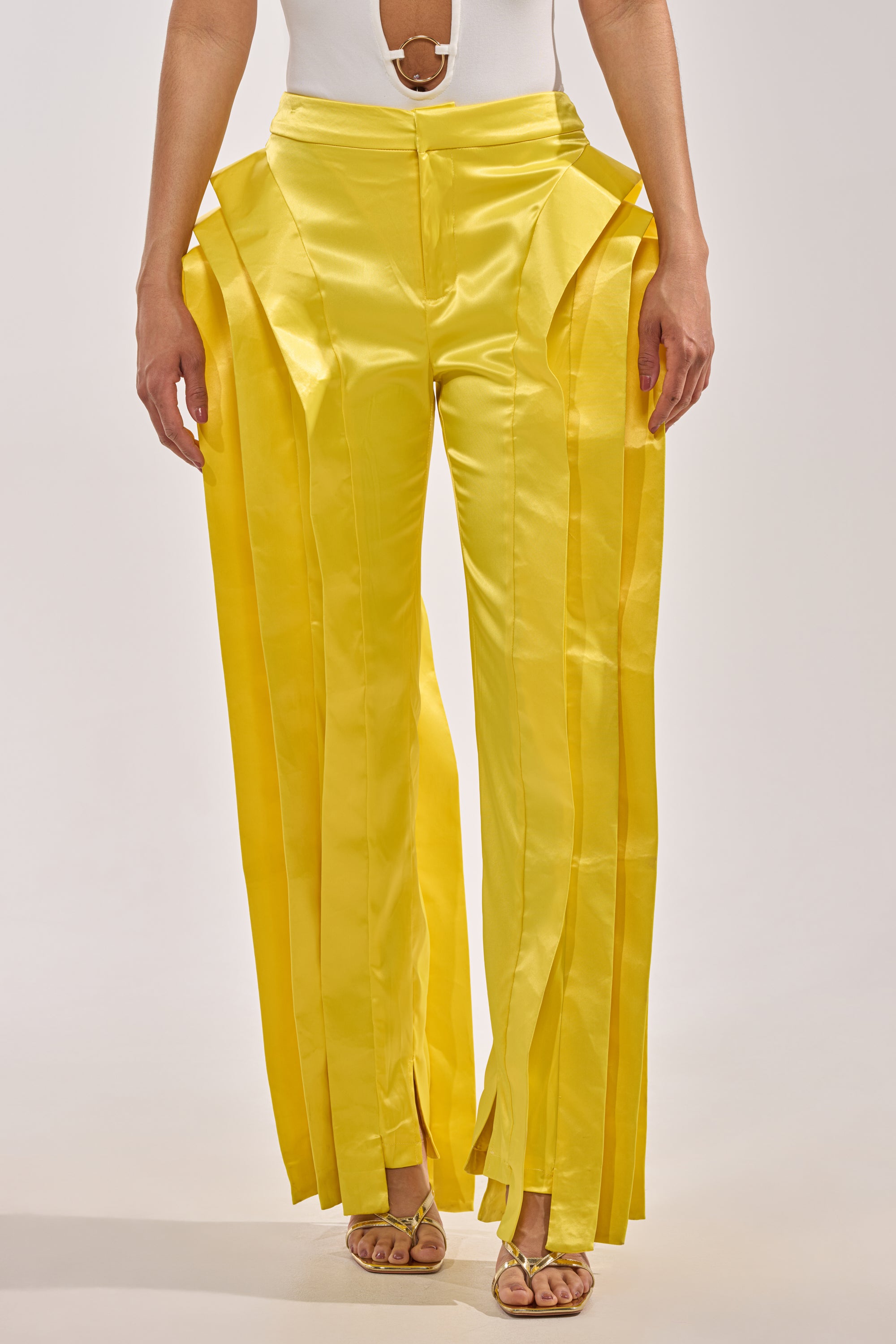 A person wearing the YELLOW INFINITY TROUSER, featuring shiny yellow satin with dramatic layered side panels and vertical pleats, styled with a white sleeveless top and open-toe sandals. 2