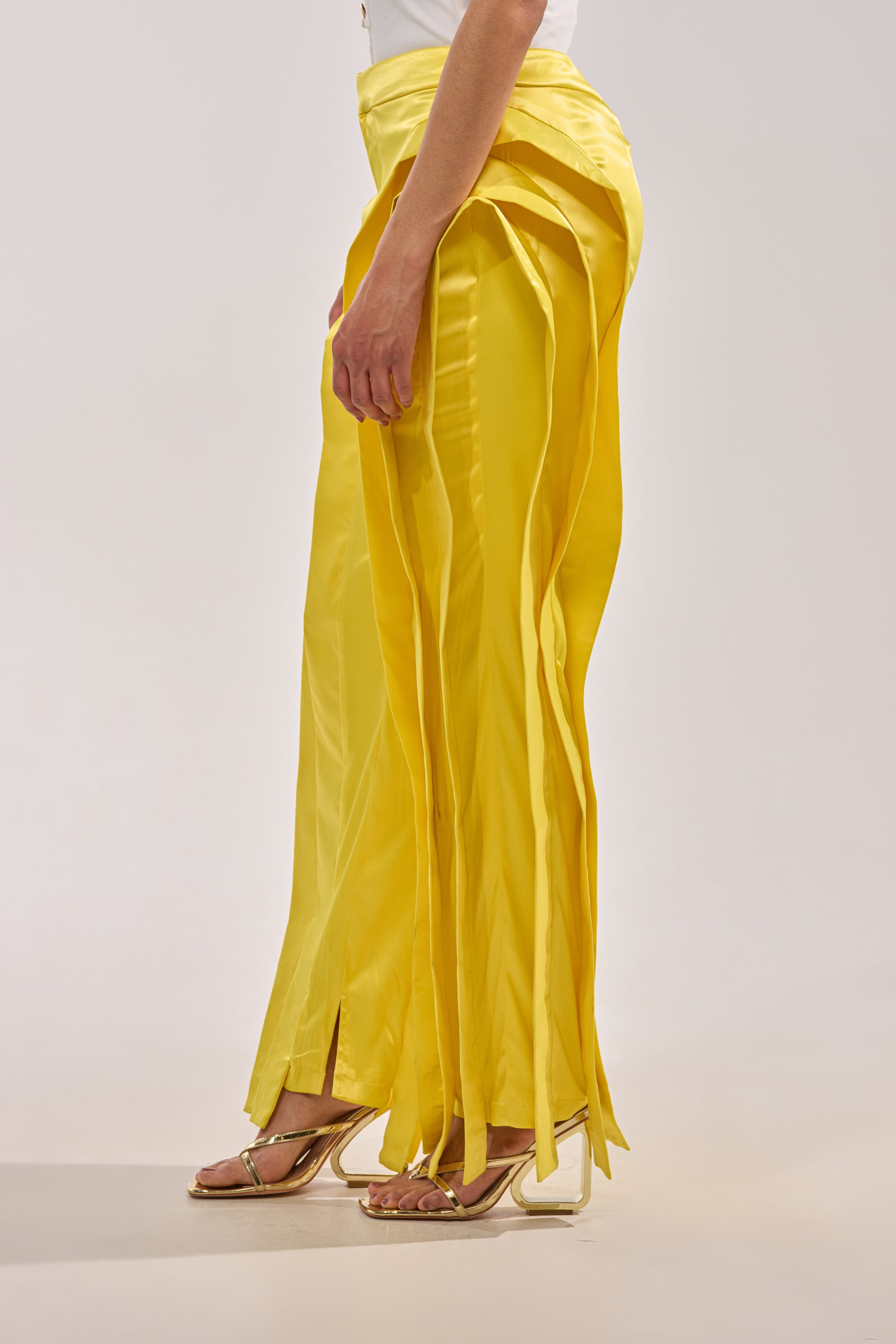 Someone poses against a plain light background wearing the YELLOW INFINITY TROUSER—bright yellow, high-rise pants with ribbon-like strips down the sides—paired with open-toe heels. 3