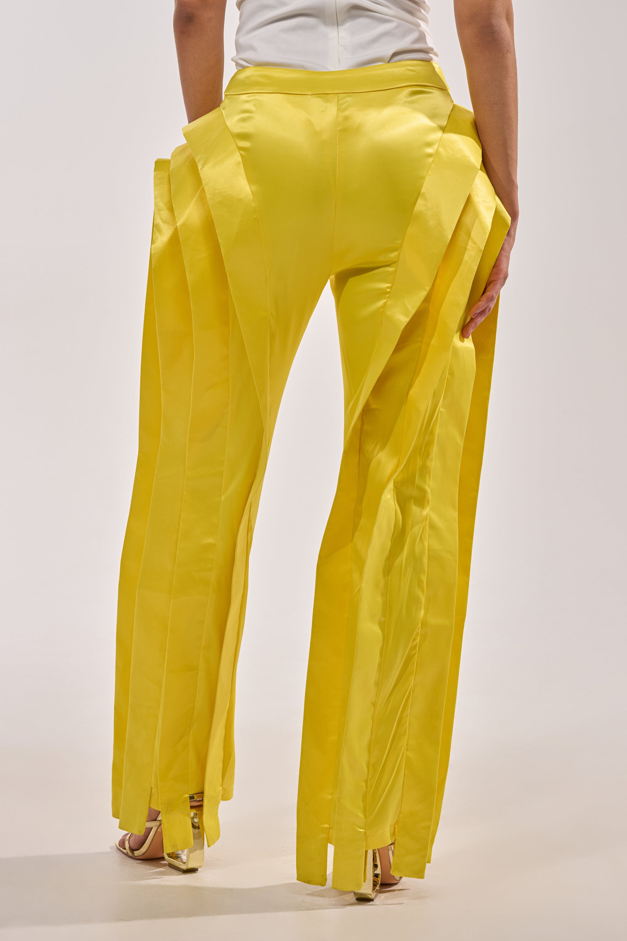 A person stands against a neutral background, shown from the back in full length, wearing the YELLOW INFINITY TROUSER—bright yellow, high-rise trousers with layered pleated panels—and a white top. 4