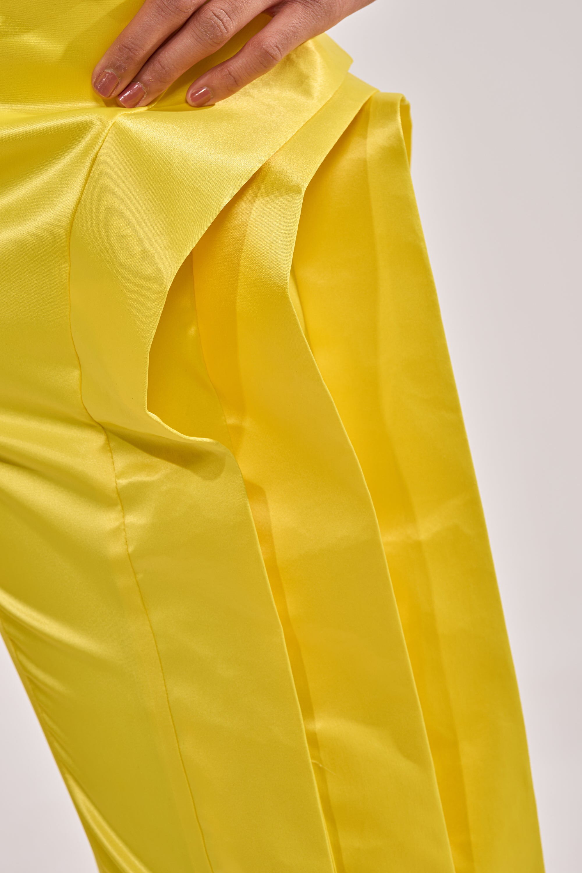 A close-up of a person wearing the YELLOW INFINITY TROUSER, featuring shiny yellow satin fabric and layered, pleated side detailing. Their hand rests near the waistband of these high-rise pants. 5