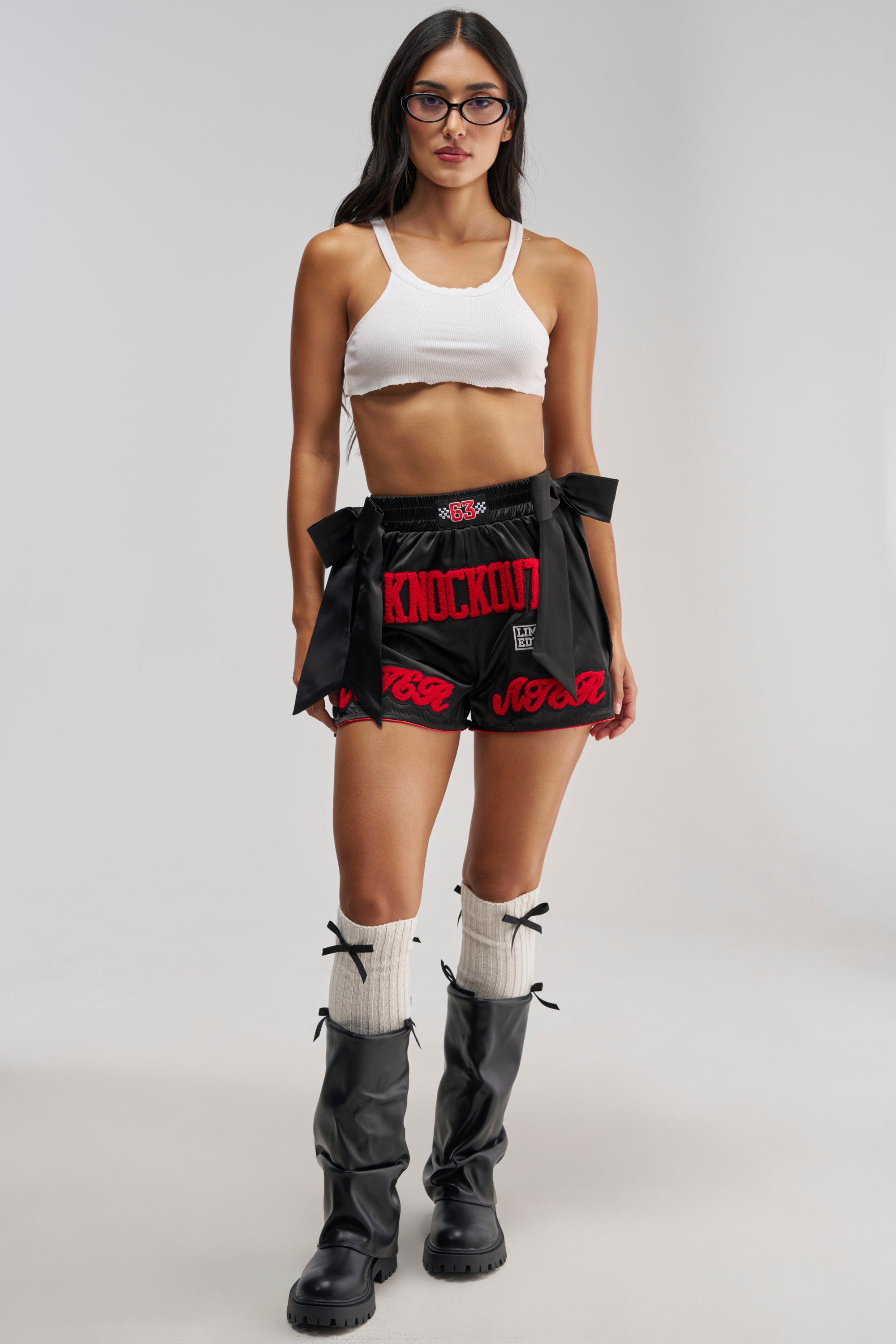 A woman stands facing forward against a plain backdrop, wearing glasses, a white crop top, white knee-high socks, black boots, and the KNOCKOUT SATIN SHORT—black satin boxer shorts featuring bold “KNOCKOUT” text.