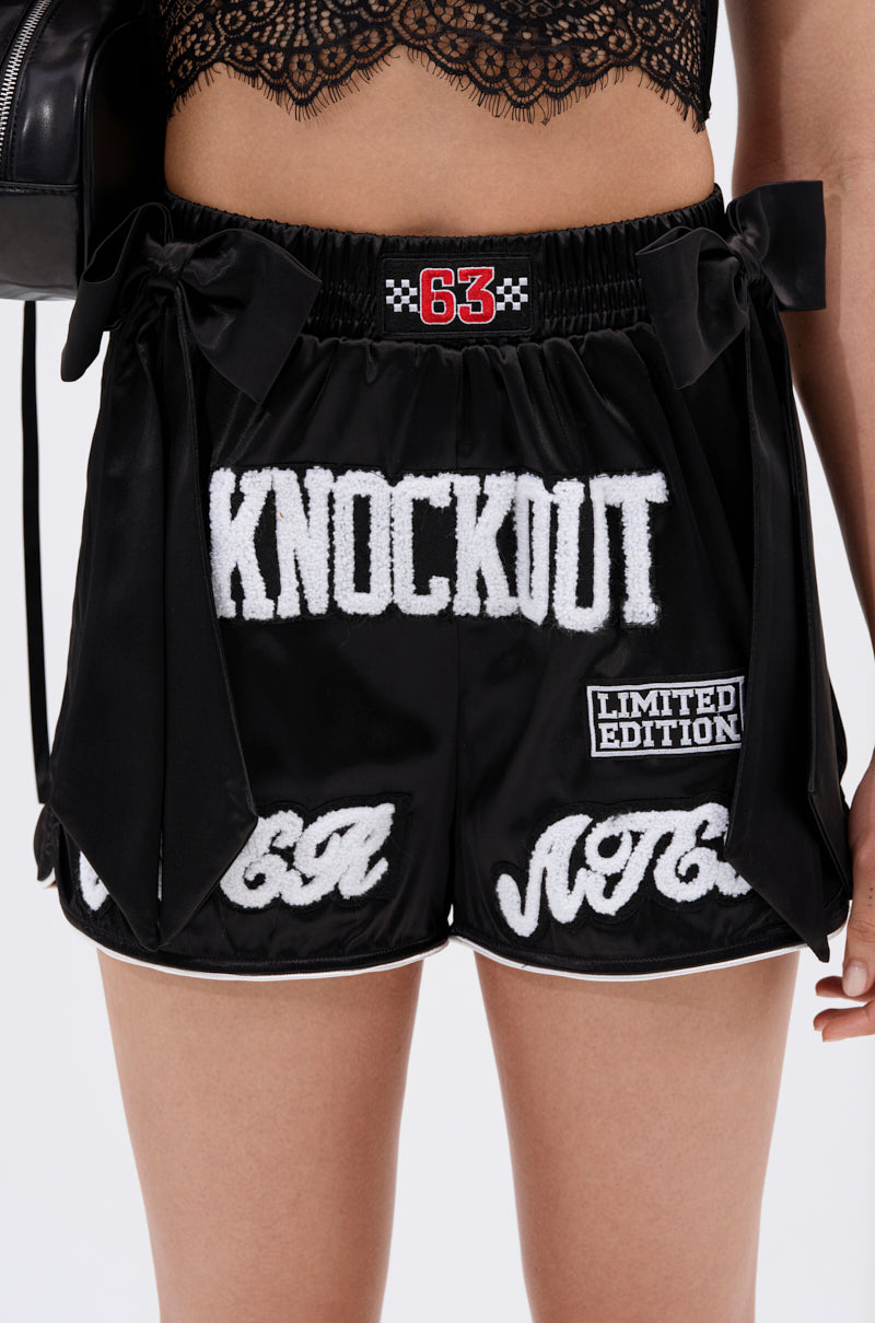 A person wears the LETS FIGHT SATIN SHORT—black, high-rise shorts with white chenille "KNOCKOUT," "LIMITED EDITION," "63," and "NOT" patches, lace trim, and side bows—paired with a black lace crop top for a bold streetwear look.