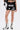 A person wears the LETS FIGHT SATIN SHORT—black satin boxing-style streetwear shorts with bold white “KNOCKOUT” text and graphic patches, high-rise fit—paired with black and white lace-up boots against a plain background.