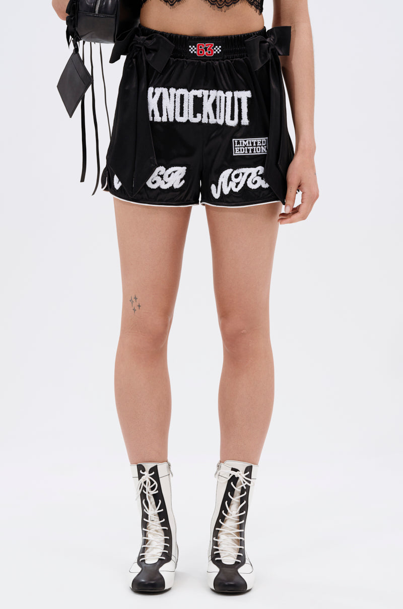 Wearing the LETS FIGHT SATIN SHORT in black with a high-rise fit and white "KNOCKOUT" lettering, styled with streetwear-inspired black and white lace-up ankle boots. Only the lower half is shown against a plain background.