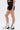 A person stands in profile wearing the LETS FIGHT SATIN SHORT—black high-rise satin shorts with a large bow—styled with a patterned crop top and tall white lace-up boots, creating a chic streetwear look against a plain background.