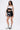 A person stands against a plain background wearing LETS FIGHT SATIN SHORTS, black and white lace-up boots, a black lace crop top under a white off-the-shoulder top, glasses, layered necklaces, and a black shoulder bag—streetwear with flair.