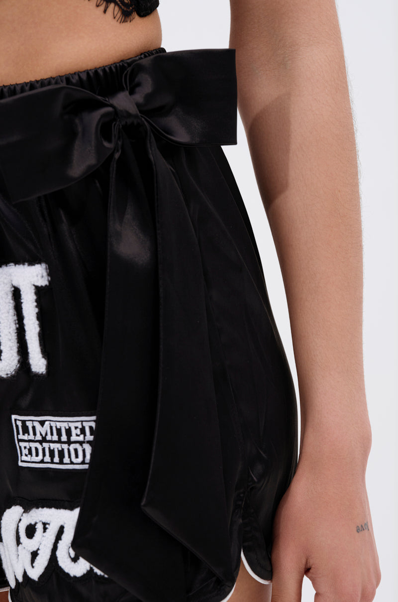 A close-up shows a person wearing the LETS FIGHT SATIN SHORT—shiny black, high-rise shorts with a large satin waist bow and several white embroidered patches, including "LIMITED EDITION." Their hand rests beside them.