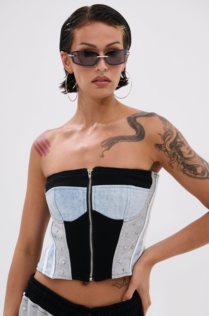 Person in the DECONSTRUCTED MASTERPIECE CORSET TOP with metallic studs, large hoop earrings, sunglasses, and visible arm and chest tattoos poses confidently against a plain light background, radiating streetwear vibe.
