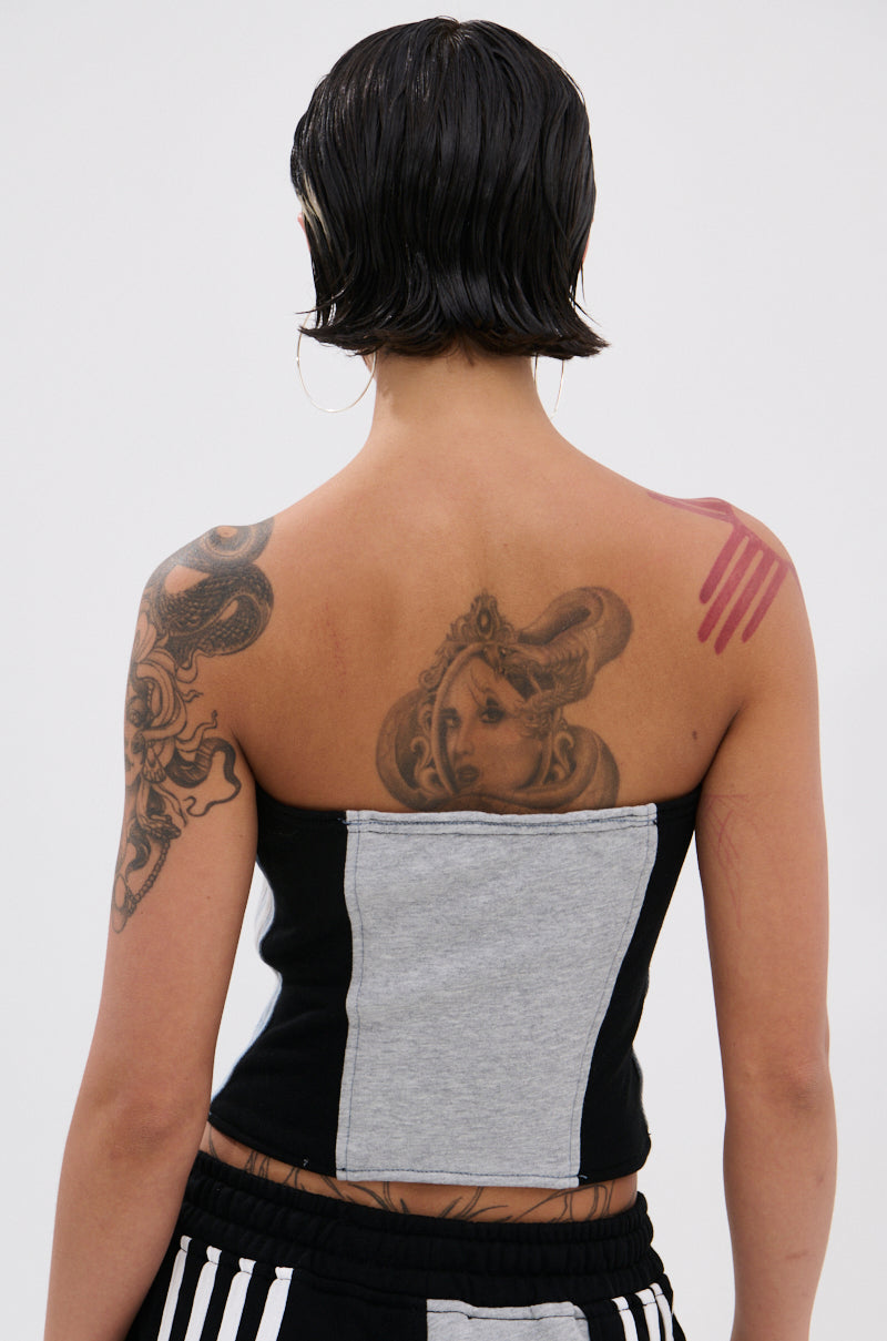 A person with short dark hair is seen from behind in a DECONSTRUCTED MASTERPIECE CORSET TOP and striped pants, showing off tattoos on their back and arms—including a portrait, abstract art, and a pink handprint—for standout streetwear style.