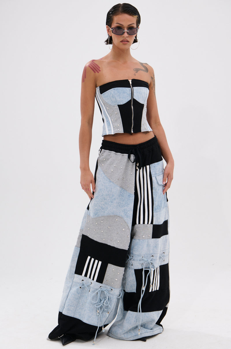 A person wears sunglasses, a DECONSTRUCTED MASTERPIECE CORSET TOP in a black and white strapless design, and wide-leg patchwork pants with streetwear flair, standing against a plain white background.