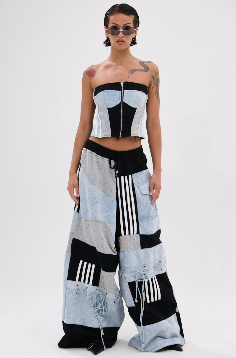 Wearing the DECONSTRUCTED MASTERPIECE CORSET TOP styled with wide-leg patchwork jeans in black, white, and denim-blue, this individual with short wet-look hair and sunglasses poses against a plain white background.