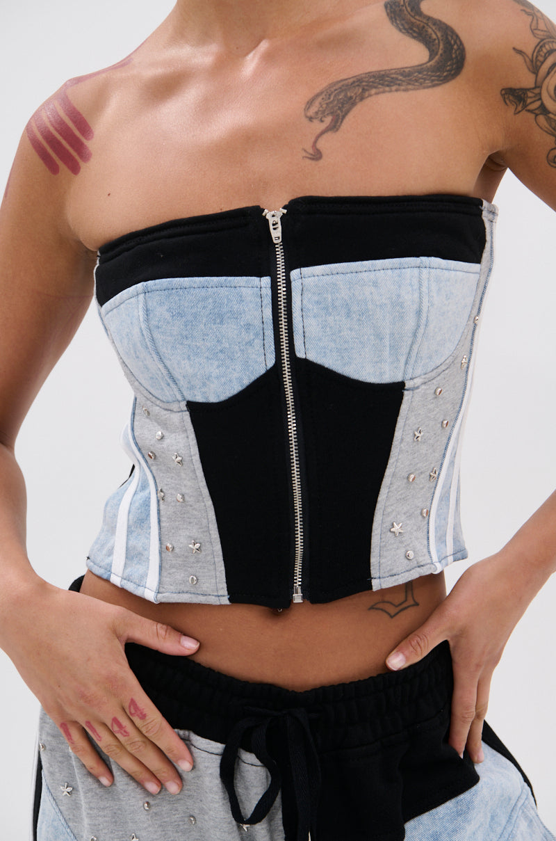 Wearing the DECONSTRUCTED MASTERPIECE CORSET TOP—a strapless denim and black zip-up with metallic studs—paired with matching sweatpants and visible tattoos, this streetwear look is captured cropped at neck and hips.