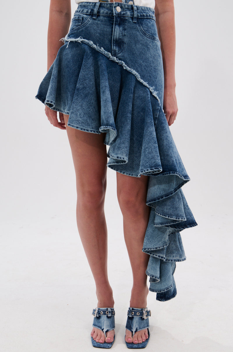 A person wears the CRAZY FOR YOU DENIM SKIRT, a high-waisted, asymmetrical blue denim skirt with ruffled, cascading high-low layers, paired with matching denim heeled sandals.