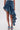A person wears the CRAZY FOR YOU DENIM SKIRT, a high-waisted, asymmetrical blue denim skirt with ruffled, cascading high-low layers, paired with matching denim heeled sandals.