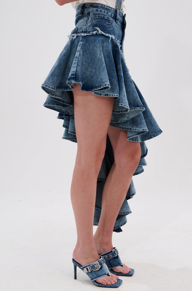 A person wears the CRAZY FOR YOU DENIM SKIRT, featuring a high-low ruffled silhouette with a distressed hem, paired with denim high-heeled sandals. The short front and layered long back stand out against a plain white background.