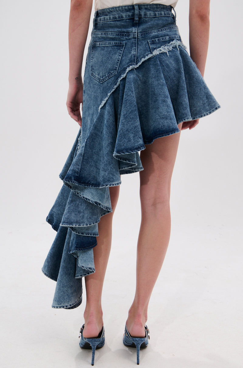 A person wears the CRAZY FOR YOU DENIM SKIRT, featuring a blue asymmetrical high-low silhouette with cascading ruffles on one side and a distressed hem on the other, perfectly paired with high-heeled shoes.