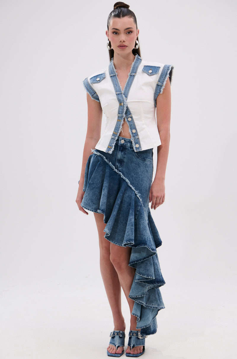 A woman models the CRAZY FOR YOU DENIM SKIRT, a high-low denim skirt with a distressed hem and dramatic ruffled detail, paired with a white sleeveless frayed denim vest and open-toe denim heels.