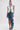 A woman models the CRAZY FOR YOU DENIM SKIRT, a high-low denim skirt with a distressed hem and dramatic ruffled detail, paired with a white sleeveless frayed denim vest and open-toe denim heels.