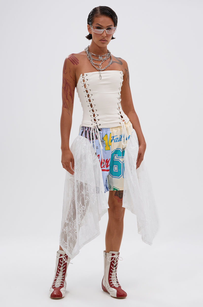A model in a white lace-up corset top, CARLY LACE GRAPHIC SHORT with colorful patchwork and lace panels, red and white lace-up boots, layered necklaces, clear glasses, and visible tattoos poses against a plain background—ideal festival streetwear. 1