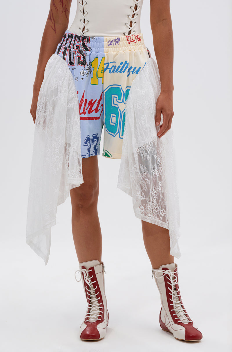 A person wears the CARLY LACE GRAPHIC SHORT, a colorful patchwork skirt with sports numbers and lace drapes, styled for festival streetwear with white and red lace-up boots and a partially visible white top, set against a plain white background. 0
