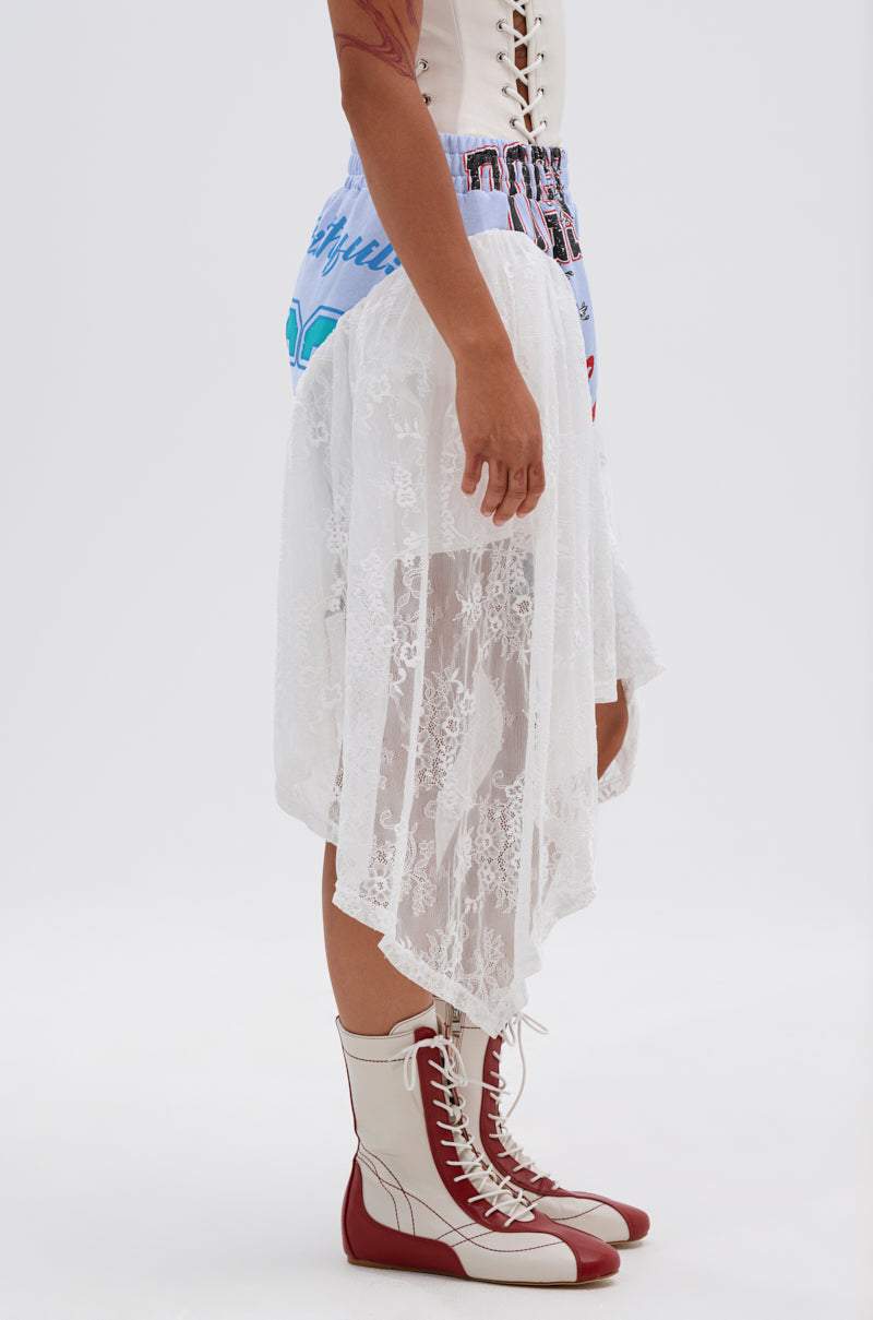 A person stands sideways in a white lace asymmetrical skirt over CARLY LACE GRAPHIC SHORT, a light corset top, and knee-high white lace-up boots with red accents. The background is plain white. 3