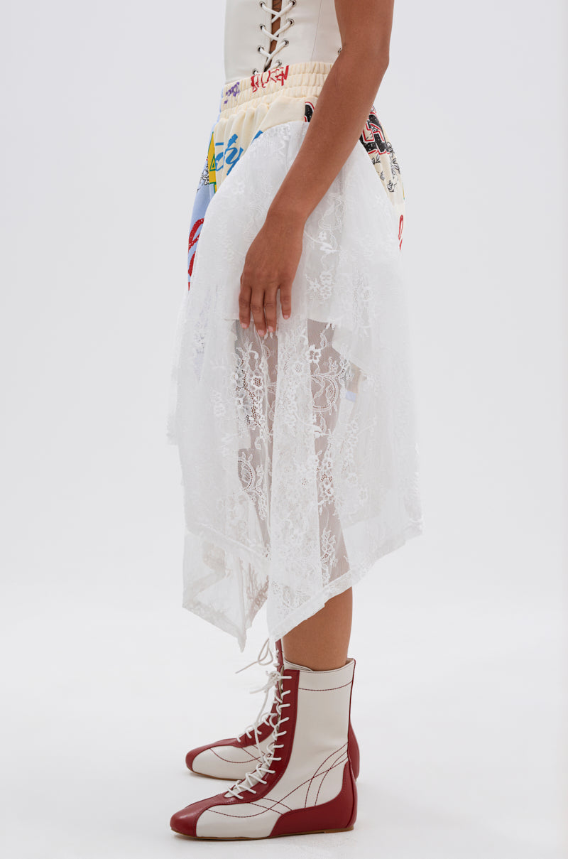 A person stands sideways wearing a white lace skirt over CARLY LACE GRAPHIC SHORTS and cream-and-red lace-up boots with pointed toes, creating a festival streetwear look with layered textures and bold graphic details. 4