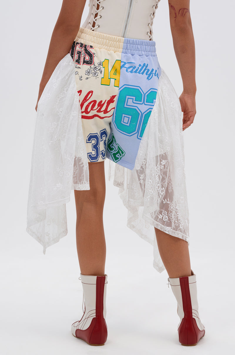 A person stands facing away, wearing the CARLY LACE GRAPHIC SHORT—patchwork high-rise shorts with colorful graphics and asymmetrical white lace—paired with a sleeveless lace-up top and white boots with red stripes. 5