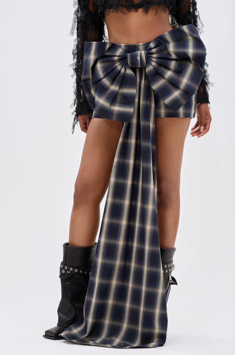 A person in black boots and a black cropped top with sheer sleeves stands wearing the PRETTY LIKE ME PLAID BOW MINI SKIRT, crafted from soft cotton-blend fabric with a long front ribbon. The background is plain and light-colored.
