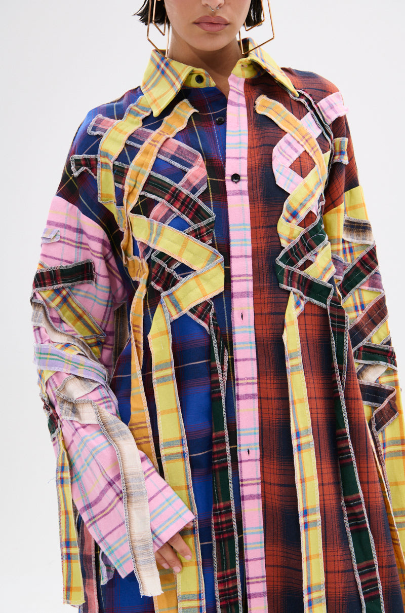 A person wears the DONT EVEN MIXED PLAID LONG BUTTON DOWN, a colorful patchwork flannel shirt dress with crisscrossed strips in yellow, pink, blue, green, and red. Its oversized fit and long sleeves give a grunge vibe. The face is partially visible.