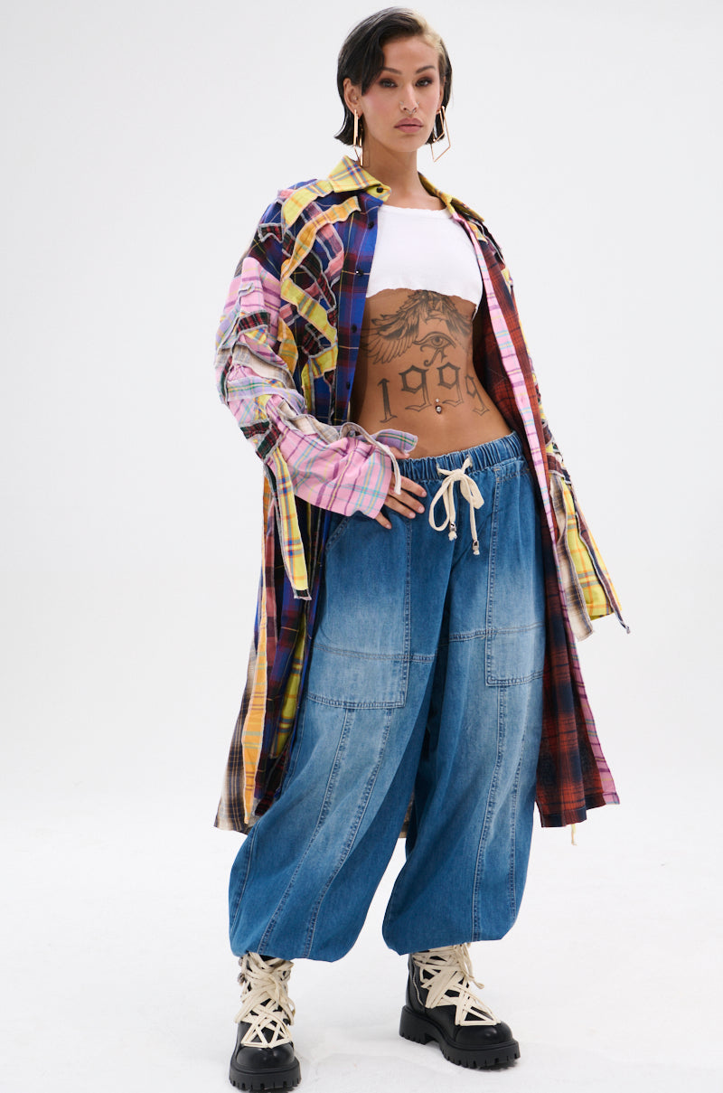 A person confidently poses in the DONT EVEN MIXED PLAID LONG BUTTON DOWN over a white crop top, paired with baggy blue jeans and chunky boots, showing off a grunge style against a plain white background.
