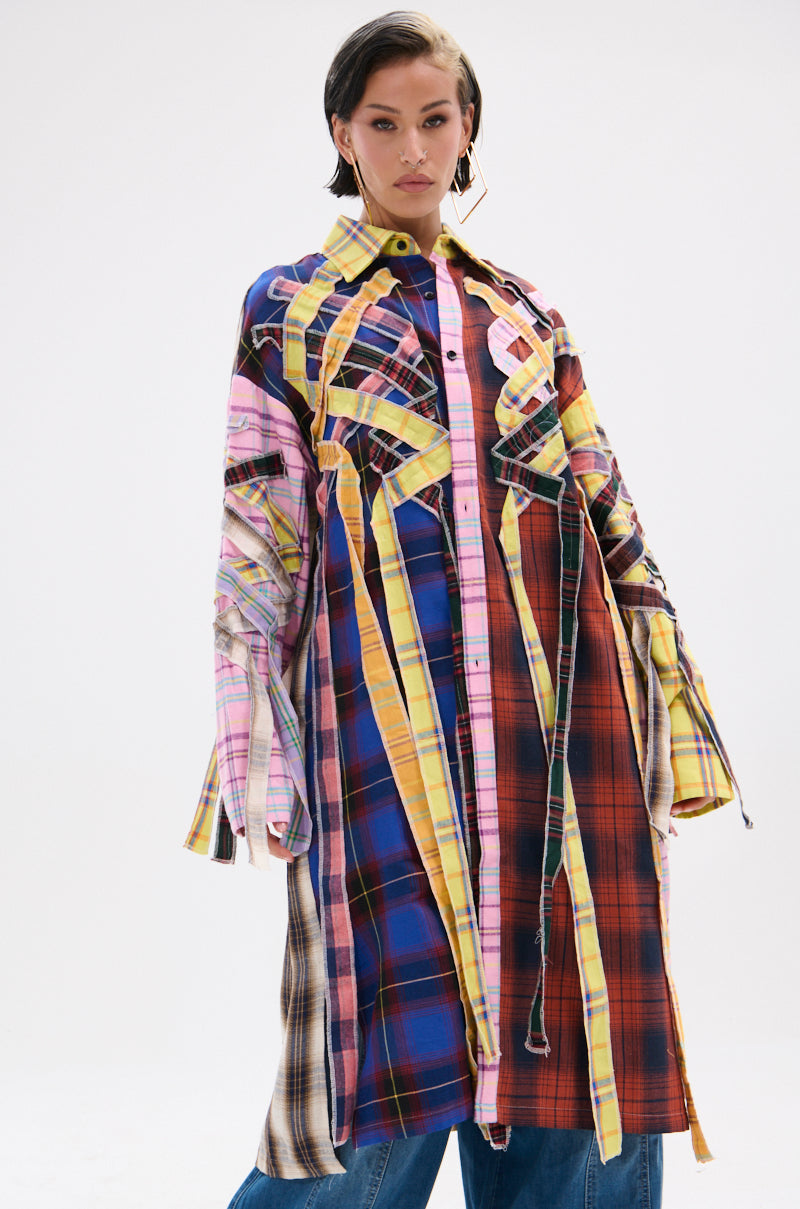 A person stands confidently in the DONT EVEN MIXED PLAID LONG BUTTON DOWN, a vibrant patchwork coat featuring bold blue, red, yellow, and pink plaid patterns, layered over denim jeans against a plain light background.