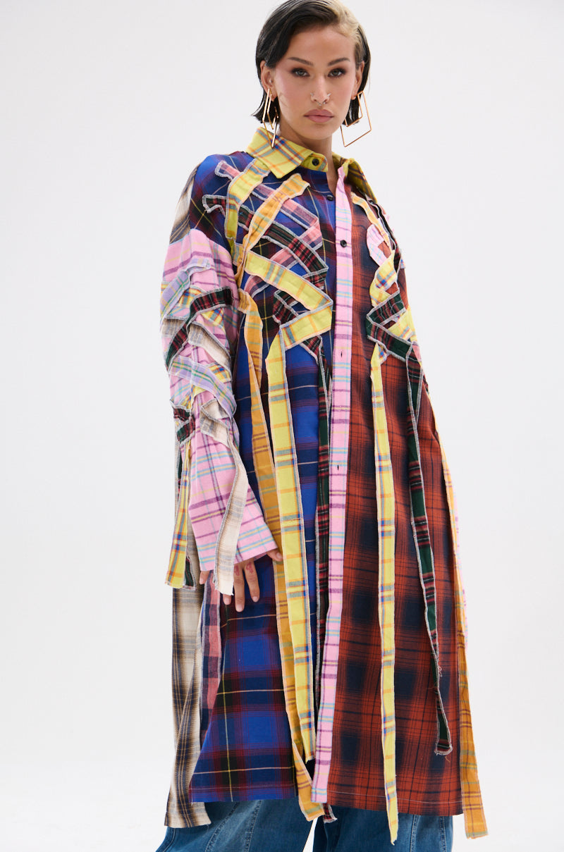 Someone wearing the DONT EVEN MIXED PLAID LONG BUTTON DOWN—a vibrant, multi-colored patchwork plaid coat in blue, pink, yellow, red, and brown—paired with blue jeans and standing against a plain white background.