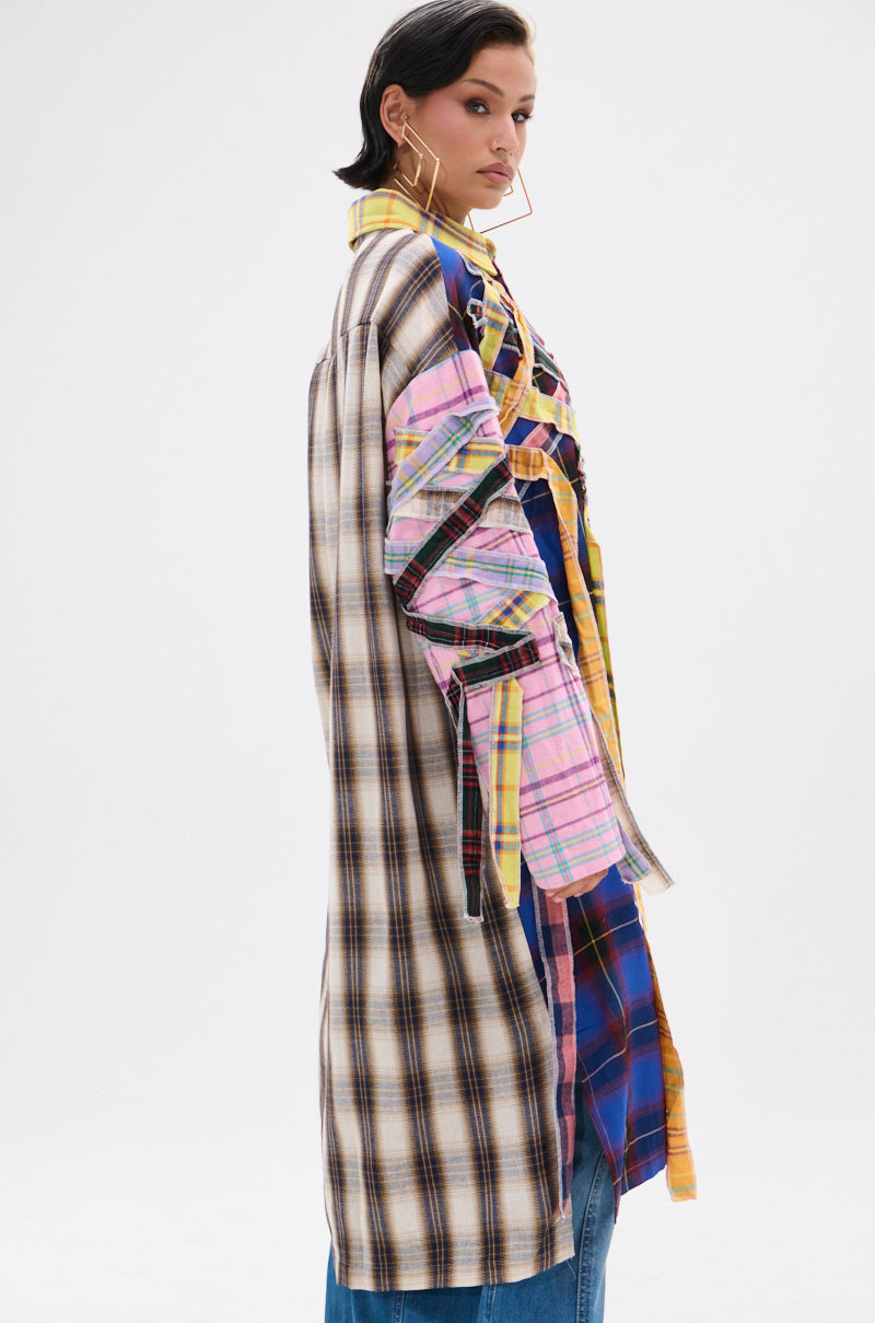 A person models the DONT EVEN MIXED PLAID LONG BUTTON DOWN—an oversized shirt with mixed check patterns—styled with blue jeans and bold geometric earrings, creating a modern grunge look against a plain backdrop.