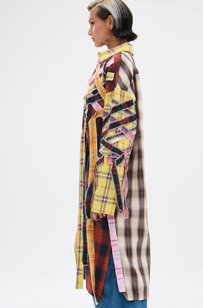 A person with short, dark hair and a blonde streak stands in profile against a white background, wearing the DONT EVEN MIXED PLAID LONG BUTTON DOWN as a grunge-style patchwork coat over blue jeans.