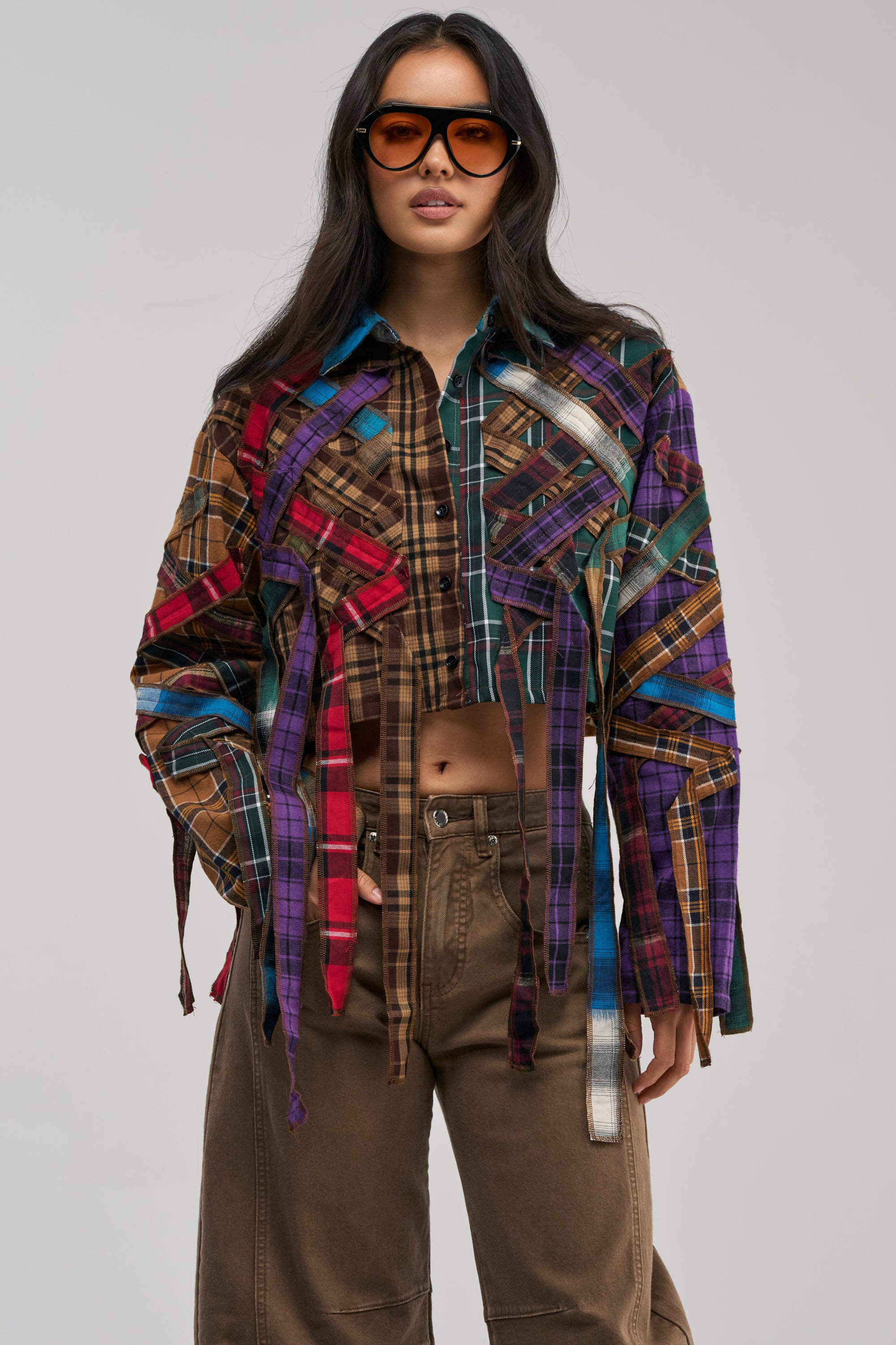 A woman wears large sunglasses, brown pants, and the CRAZY FEELING MIXED PLAID BUTTON DOWN—a multicolored plaid deconstructed lattice shirt with hanging strips. She stands facing forward against a plain light background.