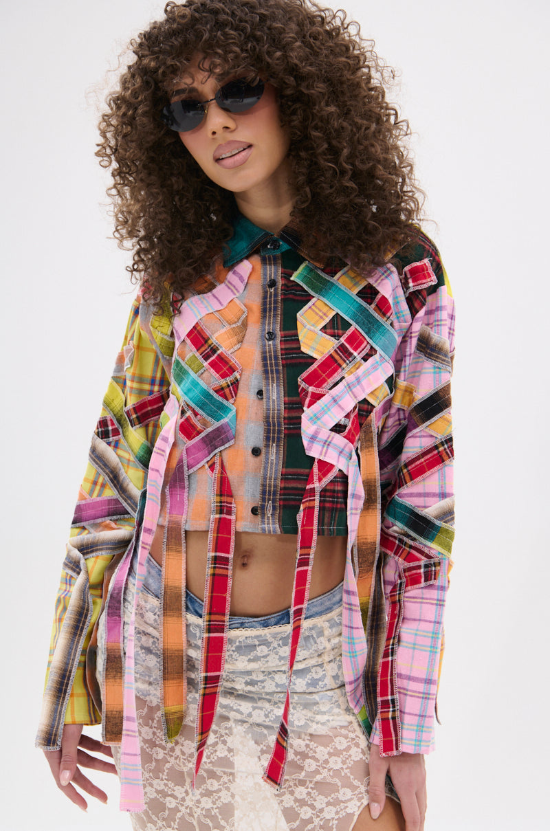 A curly-haired person in sunglasses wears the FEELING CRAZY MIXED PLAID BUTTON DOWN, a colorful punk-inspired patchwork blouse with hanging strips, over a sheer white lace skirt against a plain white background.